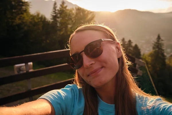 Woman in sunglasses taking selfie in mountains at sunset.