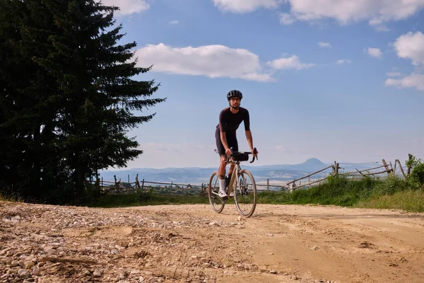 Professional cyclist practicing on gravel road.Male cyclist wearing black cycling kit and helmet.Man riding gravel bike on gravel road in mountains with scenic view.Gravel adventure.