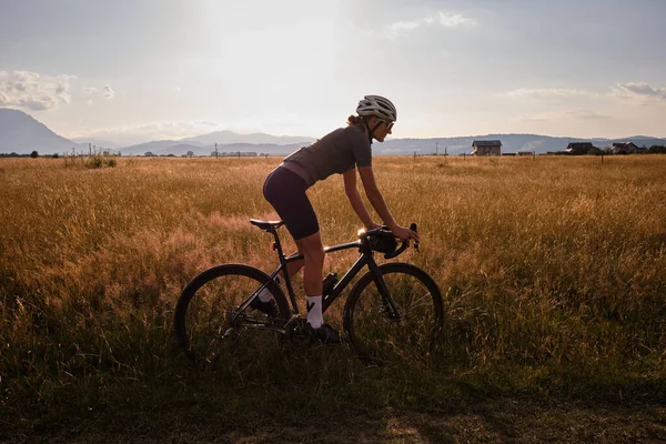 Fit female cyclist wearing cycling kit and helmet riding on the road on a gravel bike at sunset.Sports motivation image.