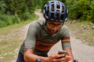 Antrenman sırasında dinlenen ve cep telefonu kullanan bir bisikletçi. Bisikletçi renkli takım elbise ve çakıl bisikletli beyaz kask giyiyor. Mobil uygulama kullanan bisikletçi.