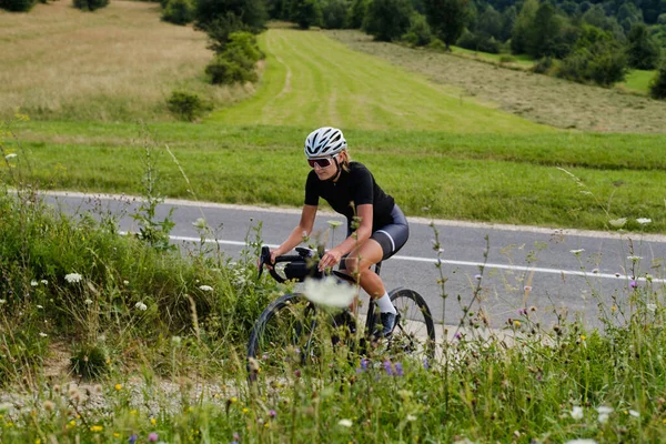 Fit female athlete wearing cycling clothing and protective helmet riding black bike with green grass around. Concept of healthy hobby and regular trainings. Gravel adventure.