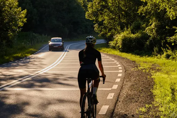 Back view of a woman cyclist in sportswear and helmet riding a black bike in the fresh air during sunset.