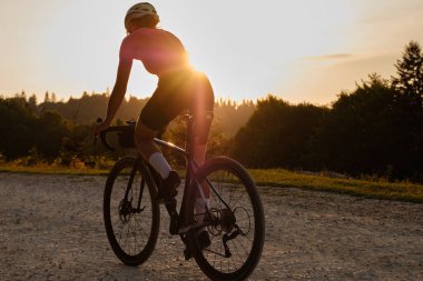 Bisiklet seti ve kask takan formda bir kadın bisikletçi gün batımında çakıllı bir yolda çakıl bisikleti sürüyor. Spor motivasyon imajı.