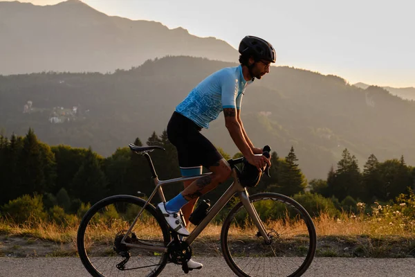 Man cyclist wearing blue cycling jersey. He is riding a gravel bike on a gravel road at sunset with a view of the mountains.Empty mountain road.Cycling gravel adventure in Romania.Bucegi Natural Park
