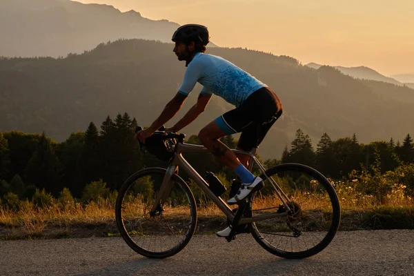 Man cyclist wearing blue cycling jersey. He is riding a gravel bike on a gravel road at sunset with a view of the mountains.Empty mountain road.Cycling gravel adventure in Romania.Bucegi Natural Park