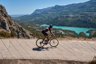 Kasklı ve bisikletli bir adam dağlık yollarda bisiklet sürüyor. Turkuaz göl manzaralı çakıl bisikletli bisikletçi. Dağlardan geçen çakıllar. Guadalest Barajı..