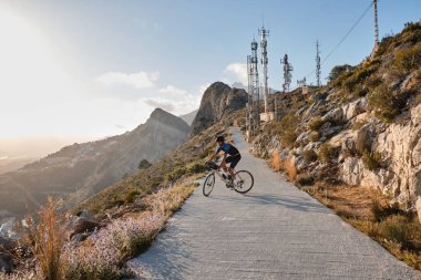 Gün batımında bisikletçinin silueti. Mavi ve siyah bisiklet seti ve çakıllı bisiklet üzerinde siyah kask takan bisikletçi arka planda dağlara doğru gidiyor. Calp, Alicante, İspanya