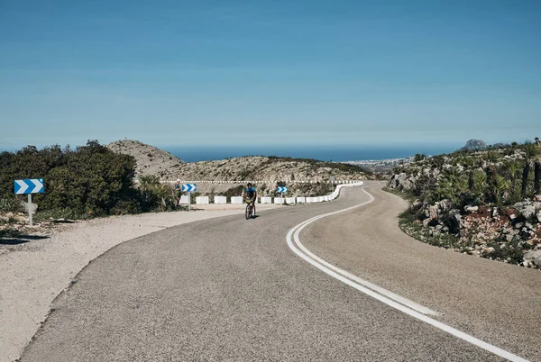 A man on a gravel bike is riding on the road in the hills with a view of the mountains.Man cyclist  wearing cycling kit and helmet.Gravel bike with cycling bag.Sport motivation.Vall de Ebo pass,Spain.