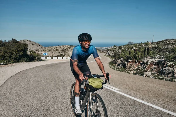 A man on a gravel bike is riding on the road in the hills with a view of the mountains.Man cyclist  wearing cycling kit and helmet.Gravel bike with cycling bag.Sport motivation.Vall de Ebo pass,Spain.