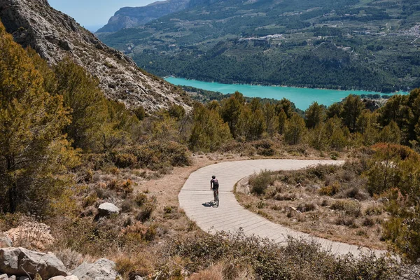 Man cyclist in helmet and cycling kit riding through mountainous roads. Cyclist on gravel bike with scenic views of turquoise lake in Spain.Gravel riding through the mountains.Guadalest Reservoir.