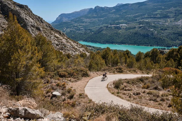 Man cyclist in helmet and cycling kit riding through mountainous roads. Cyclist on gravel bike with scenic views of turquoise lake in Spain.Gravel riding through the mountains.Guadalest Reservoir.