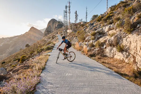 Gün batımında bisikletçinin silueti. Mavi ve siyah bisiklet seti ve çakıllı bisiklet üzerinde siyah kask takan bisikletçi arka planda dağlara doğru gidiyor. Calp, Alicante, İspanya