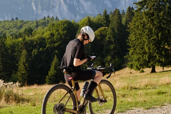 Female Cyclist Analyzes Her Ride on Gravel Bike.  Young Sportswoman and Her Gravel Bike amidst Mountain Scenery, Reviewing Achievements. Young female athlete in dark cycling kit and white helmet on gravel bike.