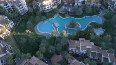Konut binalarının ve çok katlı binaların, palmiye ağaçları arasında geniş bir halka açık turkuaz havuzu olan havuzu yukarıdan aşağıya bakan görüntüsü. Torrevieja İspanya, gün batımı, İHA görüntüleri