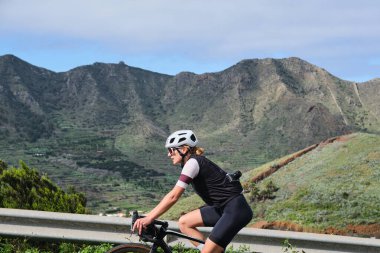 Masca Tenerife 'de virajlı dağ yolunda bisiklet süren bir kadın. Güzel bir doğada döngü. Spor giyim ve kask takan formda bir kadın bisikletçi. Spor motivasyon imajı. Tenerife 'de Bisiklet, Kanarya Adaları.