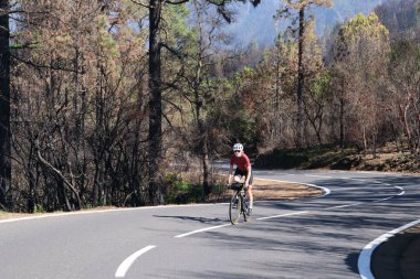 İspanya 'nın Kanarya Adaları, Tenerife' deki yanmış ormanda asfalt yolda bisiklet süren bir kadın. Spor motivasyonu. İspanya 'da bisiklet eğitimi. Bisiklet macerası. Güzel doğada pedal çevirmek..