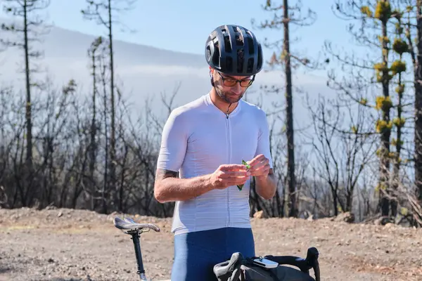Male cyclist wearing cycling kit and helmet is holding energy snack for endurance during bicycle training. Sport nutrition. Sport snack for cyclist. Granola energy bar.