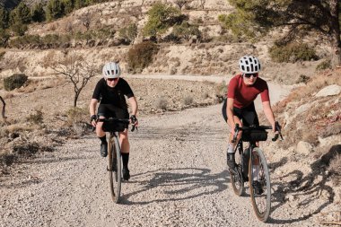 Güneşli bir yaz gününde çakıllı bir yolda çakıllı bisiklet süren iki profesyonel kadın bisikletçi. Doğal kırsalda grup bisikleti sürüşü. Çakıl gelişimi. Çakıl bisikleti macerası. Alicante, İspanya .