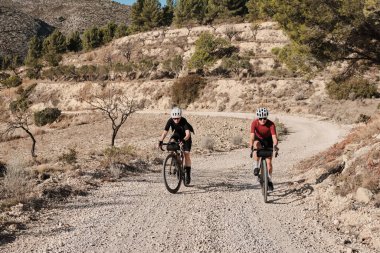 Güneşli bir yaz gününde çakıllı bir yolda çakıllı bisiklet süren iki profesyonel kadın bisikletçi. Doğal kırsalda grup bisikleti sürüşü. Çakıl gelişimi. Çakıl bisikleti macerası. Alicante, İspanya .