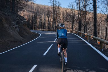 İspanya 'nın Kanarya Adaları, Tenerife' deki yanmış ormanın içinden asfalt yolda giden kadın bisikletçi. Spor motivasyonu. İspanya 'da bisiklet eğitimi. Hippi kadın bisikletçi. Bisiklet macerası .