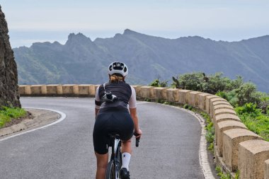 Masca Tenerife 'de virajlı dağ yolunda bisiklet süren bir kadın. Güzel bir doğada döngü. Spor giyim ve kask takan formda bir kadın bisikletçi. Spor motivasyon imajı. Tenerife 'de Bisiklet, Kanarya Adaları.