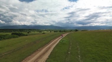 Dağ ve yeşil tarlalarla çevrili inanılmaz destansı bir manzarada bisiklet süren kadın bisikletçiyi takip eden drone. Doğal parktaki asfaltsız yolda çakıl bisikleti sürüyor. Çakıl bisikleti konsepti.