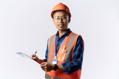 Güvenlik teçhizatı takan Asyalı erkek inşaat işçisi elinde bir dosya, saha müfettişi, endüstriyel ve inşaat konsepti, izole edilmiş beyaz arka plan taşırken kameraya güvenle bakıyor..
