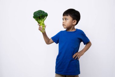 Asian boy in blue shirt looks very excited and confident standing while posing with broccoli vegetable, concept of healthy lifestyle, isolated in white background. clipart