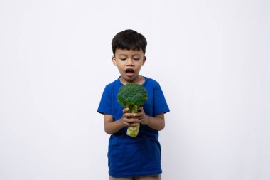 Asian boy in blue shirt looks very excited and confident standing while posing with broccoli vegetable, concept of healthy lifestyle, isolated in white background. clipart