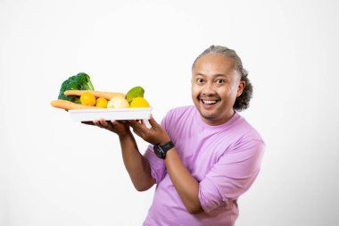 Kıvırcık saçlı, yetişkin bir Asyalı erkek, bir tepsi taze sebze taşırken onu işaret ediyor, sağlıklı ve besleyici taze sebze yeme kampanyası, sağlıklı yaşam tarzı