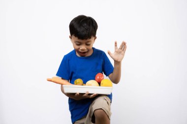 a young Asian boy in a vibrant blue shirt standing confidently with a tray full of assorted vegetables and fruits, concept of a healthy lifestyle, isolated white background. clipart