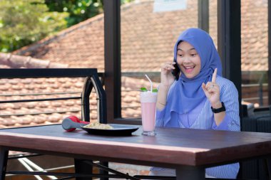 Genç Müslüman kadın tesettürlü, kafede oturmuş meyve suyunun, atıştırmanın ve kahvenin tadını çıkarıyor. Telefon, kırmızı kulaklık, müzik.