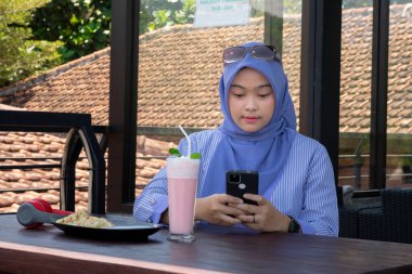 Genç Müslüman kadın tesettürlü, kafede oturmuş meyve suyunun, atıştırmanın ve kahvenin tadını çıkarıyor. Telefon, kırmızı kulaklık, müzik.