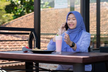 Genç Müslüman kadın tesettürlü, kafede oturmuş meyve suyunun, atıştırmanın ve kahvenin tadını çıkarıyor. Telefon, kırmızı kulaklık, müzik.