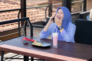 Genç Müslüman kadın tesettürlü, kafede oturmuş meyve suyunun, atıştırmanın ve kahvenin tadını çıkarıyor. Telefon, kırmızı kulaklık, müzik.