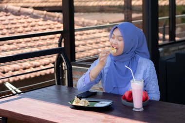 Genç Müslüman kadın tesettürlü, kafede oturmuş meyve suyunun, atıştırmanın ve kahvenin tadını çıkarıyor. Telefon, kırmızı kulaklık, müzik.