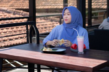 Genç Müslüman kadın tesettürlü, kafede oturmuş meyve suyunun, atıştırmanın ve kahvenin tadını çıkarıyor. Telefon, kırmızı kulaklık, müzik.