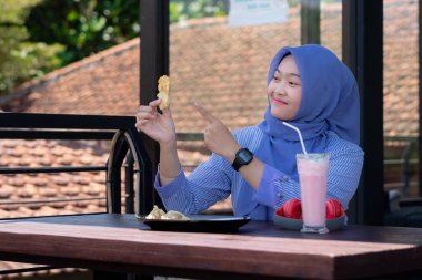 Genç Müslüman kadın tesettürlü, kafede oturmuş meyve suyunun, atıştırmanın ve kahvenin tadını çıkarıyor. Telefon, kırmızı kulaklık, müzik.