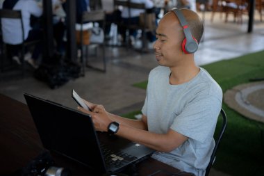 Kel bir adam kırmızı bir telefon ve kırmızı bir kulaklıkla masada dizüstü bilgisayarı olan bir kahve dükkanının terasında gülümsüyor ve konuşuyor.