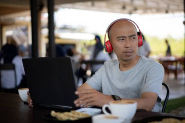 Kel bir adam kırmızı bir telefon ve kırmızı bir kulaklıkla masada dizüstü bilgisayarı olan bir kahve dükkanının terasında gülümsüyor ve konuşuyor.