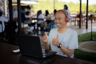 Kel bir adam kırmızı bir telefon ve kırmızı bir kulaklıkla masada dizüstü bilgisayarı olan bir kahve dükkanının terasında gülümsüyor ve konuşuyor.