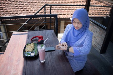 Başörtüsü takan genç bir Asyalı kadın arkadaşının kafeye gelmesini beklerken saatine bakarken çok üzgün ve hayal kırıklığına uğramış görünüyor..