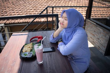 Başörtüsü takan genç bir Asyalı kadın arkadaşının kafeye gelmesini beklerken saatine bakarken çok üzgün ve hayal kırıklığına uğramış görünüyor..