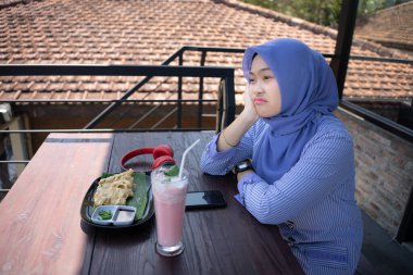 Başörtüsü takan genç bir Asyalı kadın arkadaşının kafeye gelmesini beklerken saatine bakarken çok üzgün ve hayal kırıklığına uğramış görünüyor..