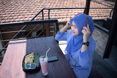 Genç Asyalı tesettürlü bir kadın, bir kafede dinlenirken cep telefonundan bir müzik çalarıyla müzik dinlemekten zevk alıyor. Teknoloji konsepti..