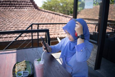 Genç Asyalı tesettürlü bir kadın, bir kafede dinlenirken cep telefonundan bir müzik çalarıyla müzik dinlemekten zevk alıyor. Teknoloji konsepti..