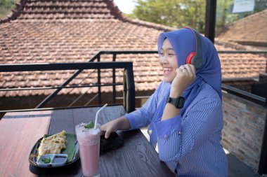 Genç Asyalı tesettürlü bir kadın, bir kafede dinlenirken cep telefonundan bir müzik çalarıyla müzik dinlemekten zevk alıyor. Teknoloji konsepti..