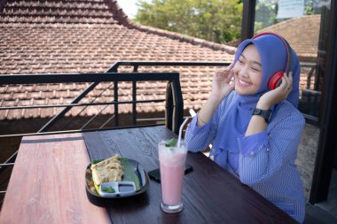 Genç Asyalı tesettürlü bir kadın, bir kafede dinlenirken cep telefonundan bir müzik çalarıyla müzik dinlemekten zevk alıyor. Teknoloji konsepti..