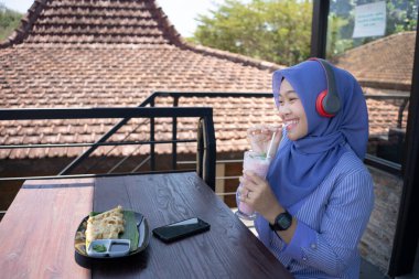 Genç Asyalı tesettürlü bir kadın, bir kafede dinlenirken cep telefonundan bir müzik çalarıyla müzik dinlemekten zevk alıyor. Teknoloji konsepti..