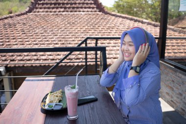 Genç Asyalı tesettürlü bir kadın, bir kafede dinlenirken cep telefonundan bir müzik çalarıyla müzik dinlemekten zevk alıyor. Teknoloji konsepti..